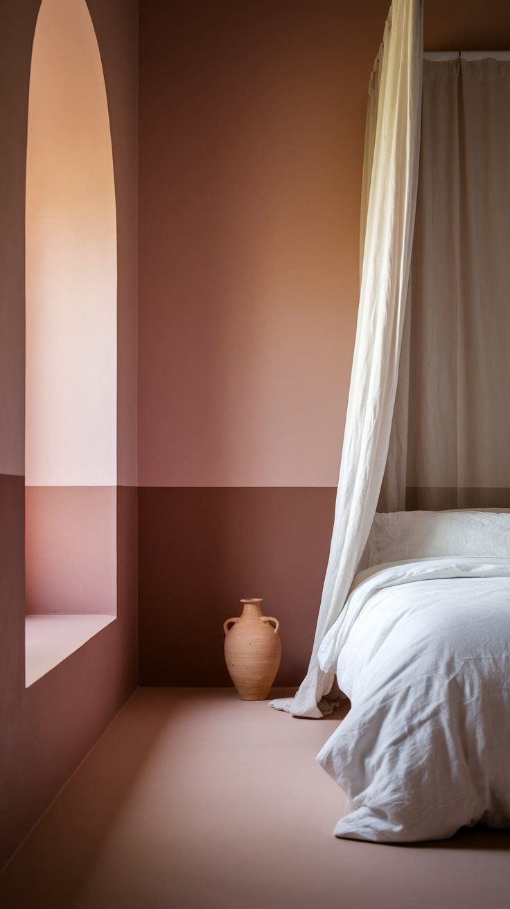 A minimalist bedroom features soft two-tone pink walls and a matching arched niche beside a bed draped in flowing white linens. A simple terracotta urn rests on the floor, adding an earthy accent to the clean, modern space.